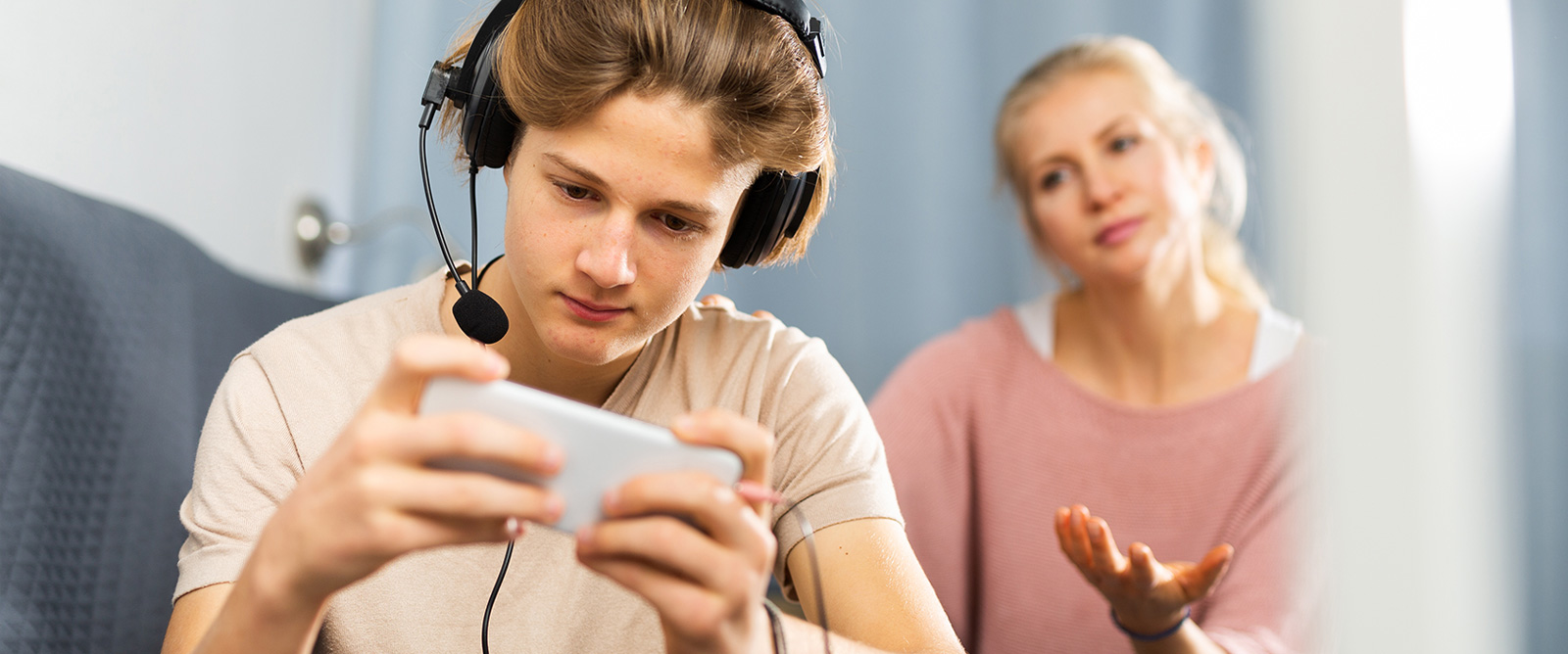 Tonårskille sitter med hörlurar och telefon, i bakgrunden hans mamma som inte når fram.